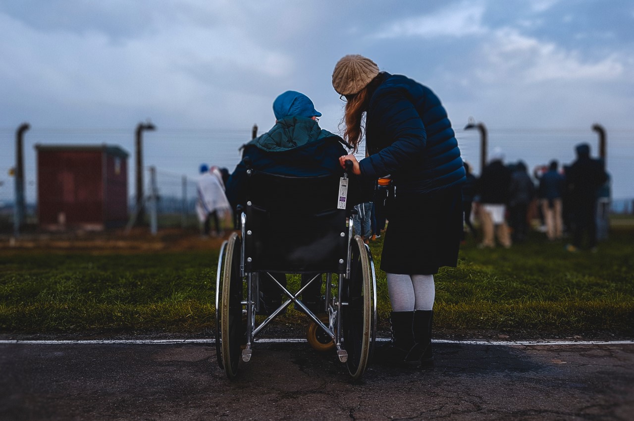 A lady outside in a wheelchair, with her friend at her side, looking towards a crowd of people all watching something through a fence - Trauma Insurance