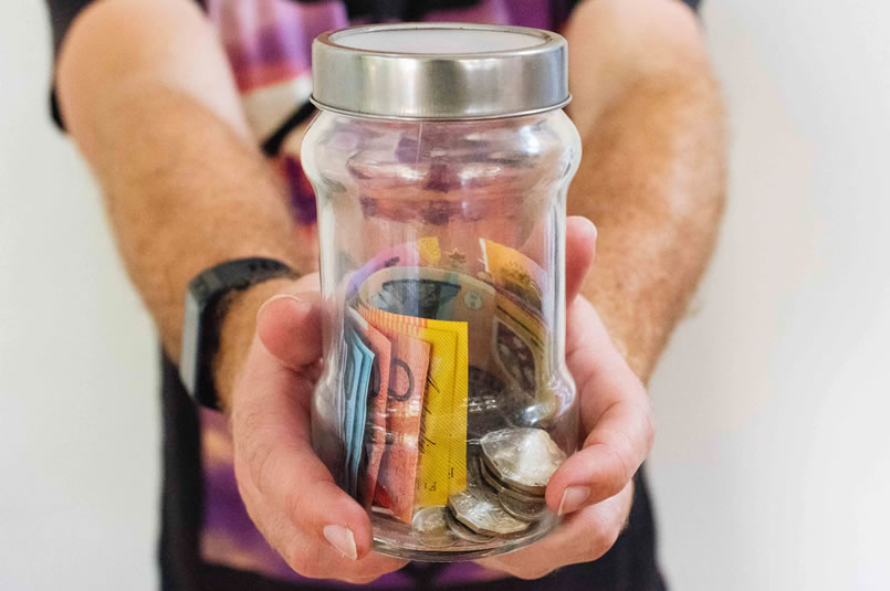 Ways to Save Money Around the Home: A man hold out a large glass jar filled with a mixture of Australian dollar notes and coins.