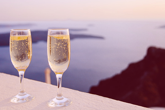Two glasses of bubbling champagne on a ledge, in the evening sun, with sea views in the background. - Baldwin Financial Services | Prospect Financial Advisors.