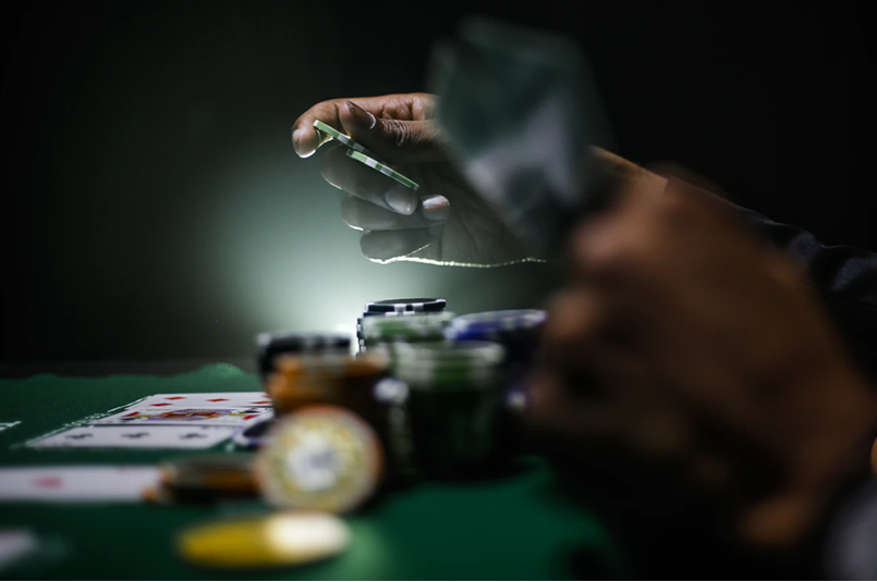 Investing Versus Gambling: The hands of a person playing poker in a dark room, with cards and poker chips on the table in front of him.