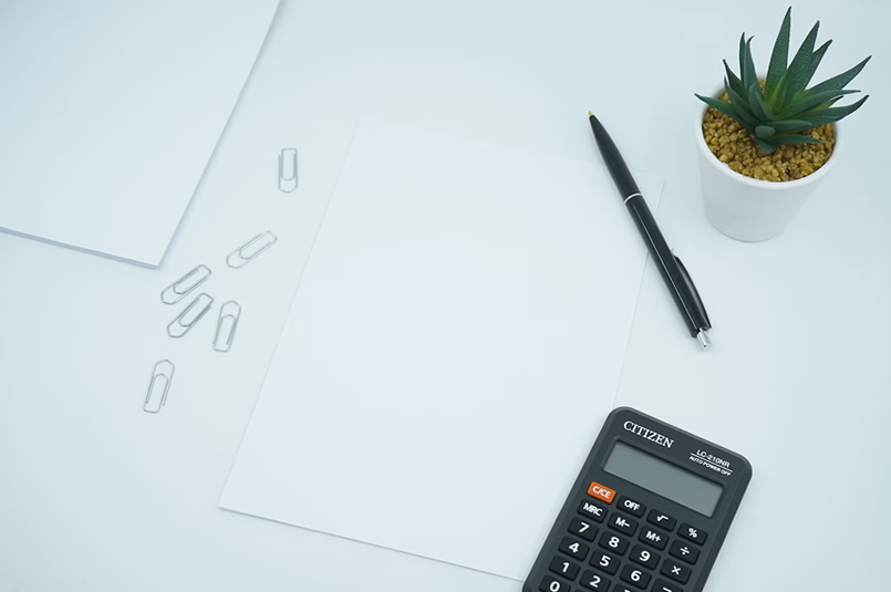 A calculator, a pen, paperclips and several sheets of blank paper on top of a white desk.